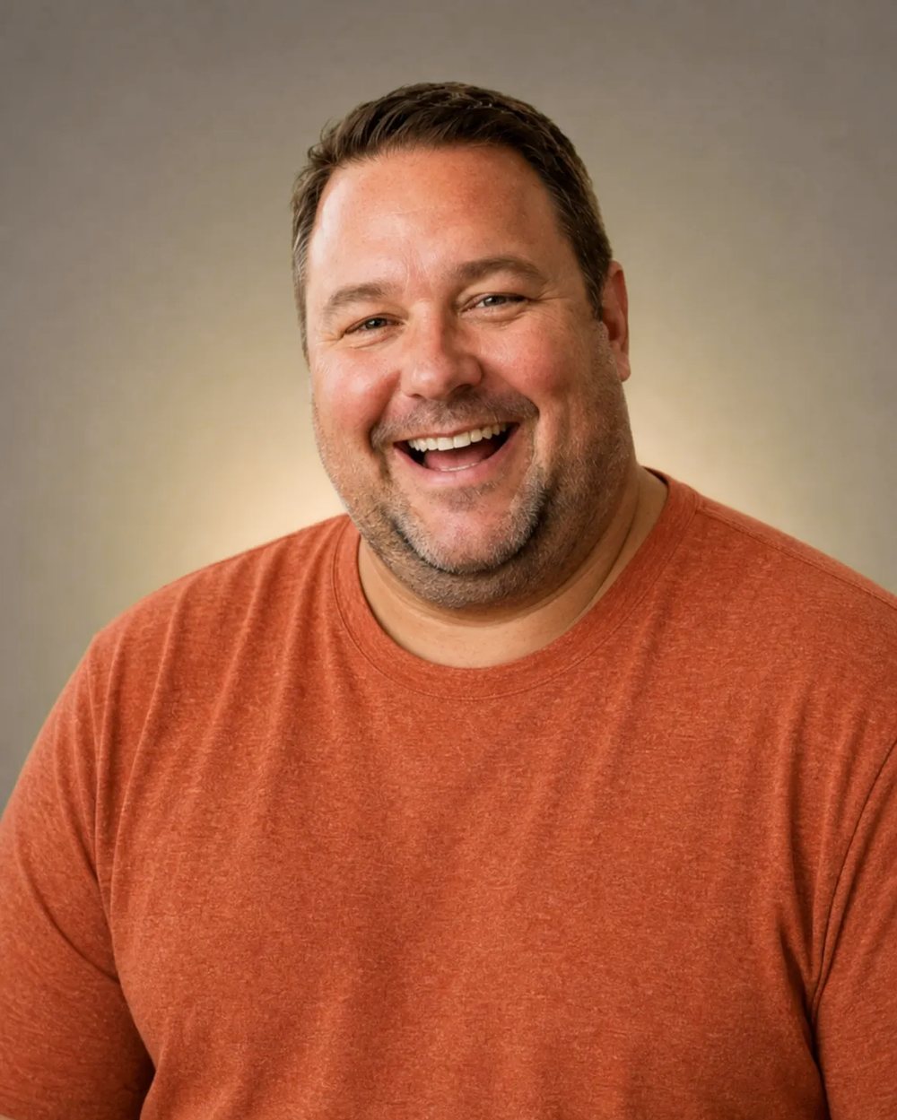 A man laughing in a rust-coloured tee against a warm neutral backdrop
